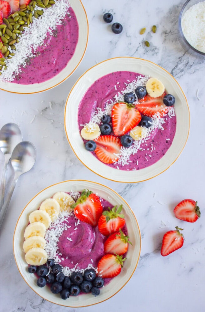 Three smoothie bowls topped with sliced strawberries, bananas, blueberries, grated coconut, and seeds, placed on a marble surface with scattered fruit and two spoons nearby.