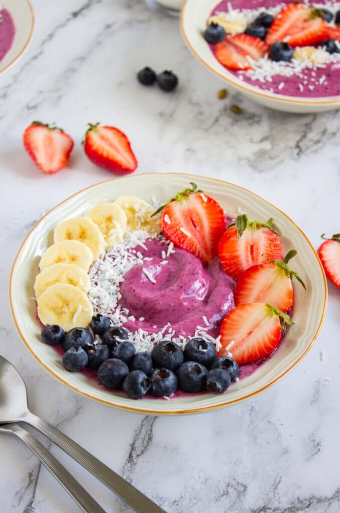A smoothie bowl topped with sliced bananas, strawberries, blueberries, and shredded coconut, placed on a marble surface with scattered berries and two spoons nearby.