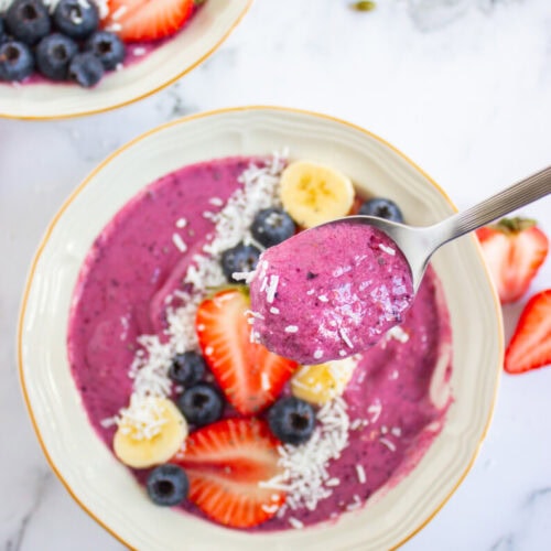 A bowl of purple smoothie topped with blueberries, banana slices, strawberries, and shredded coconut, with a spoonful held above it. Another similar bowl is partially visible.