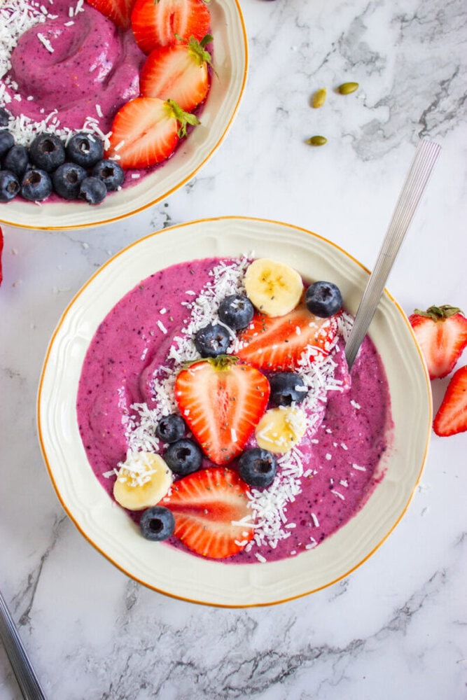 Smoothie bowl with purple mixture topped with sliced strawberries, blueberries, banana slices, shredded coconut, and seeds, accompanied by a spoon on the side.