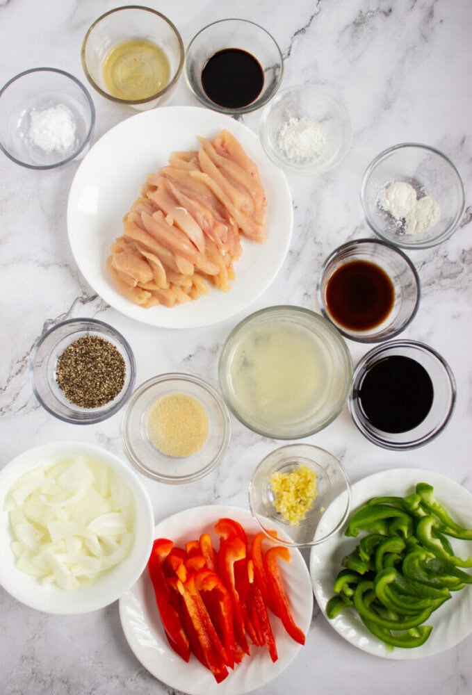 Raw chicken strips, sliced onions, bell peppers, and various seasonings and sauces arranged in glass bowls on a marble surface.