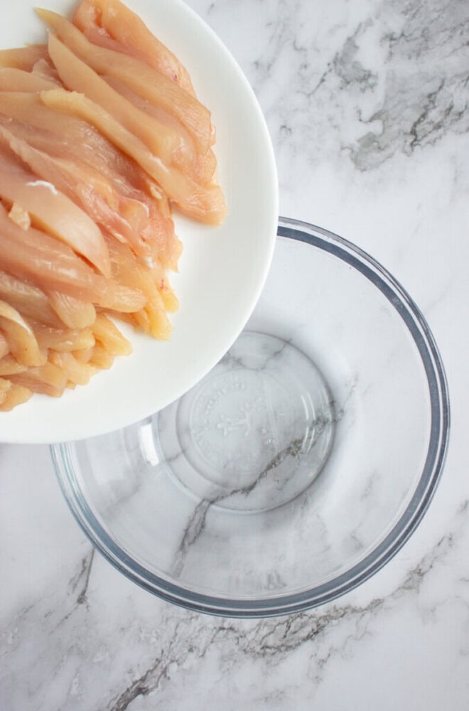 Sliced raw chicken breast is being transferred from a white plate into an empty clear glass bowl on a marble countertop.
