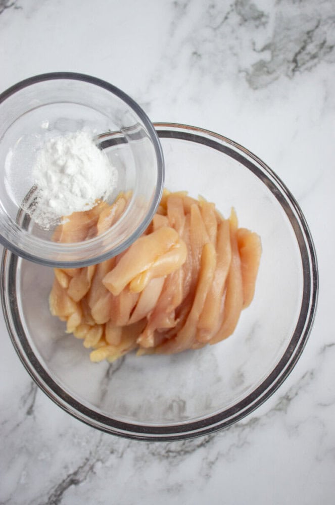 A bowl of raw, sliced chicken breast with a small bowl of white powder, likely cornstarch, positioned above it on a marble surface.