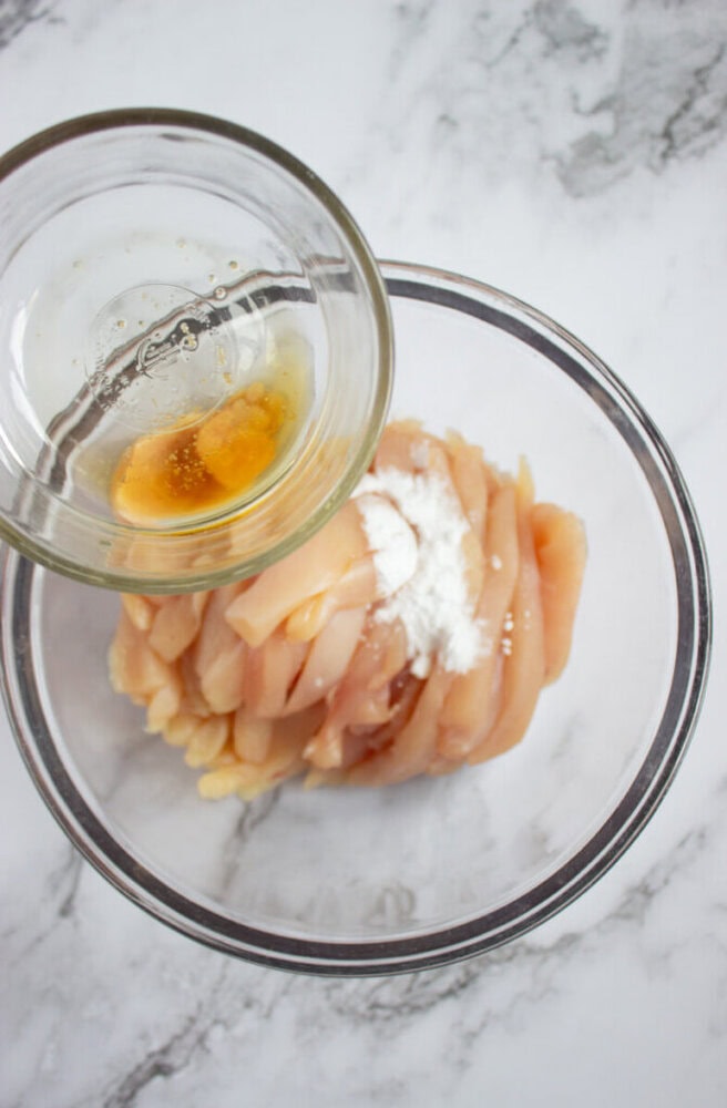 A glass bowl of sliced raw chicken with white powder, as liquid from a small bowl is poured over it, on a marble countertop.