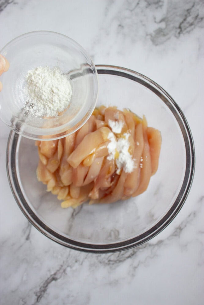 A glass bowl with raw sliced chicken breast, some white powder on top, and a hand adding more white powder from a small bowl on a marble surface.