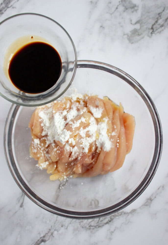 A glass bowl of raw sliced chicken topped with white cornstarch, next to a small bowl of dark soy sauce on a marble surface.