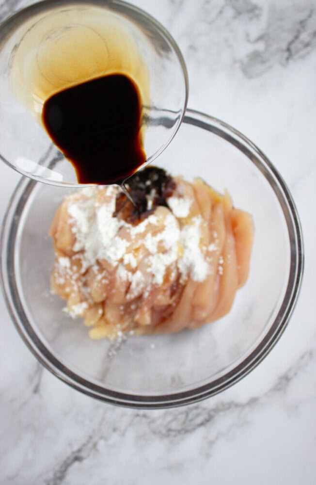 A glass bowl with raw sliced chicken, cornstarch, and minced garlic, while soy sauce is being poured over the top.
