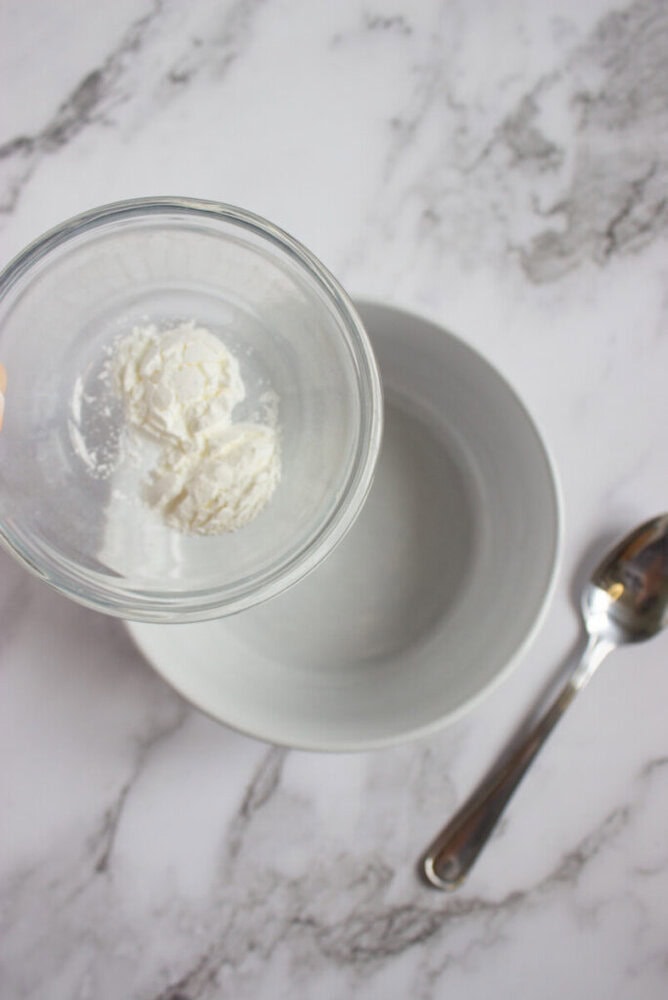 A hand holds a glass bowl with white powder over an empty gray bowl, next to a metal spoon on a marble countertop.