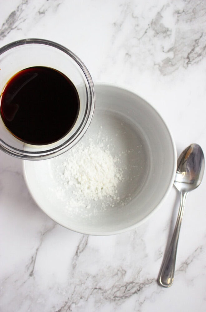 A bowl with white powder next to a spoon on a marble surface; a hand holds a cup of dark liquid above the bowl, about to pour it in.