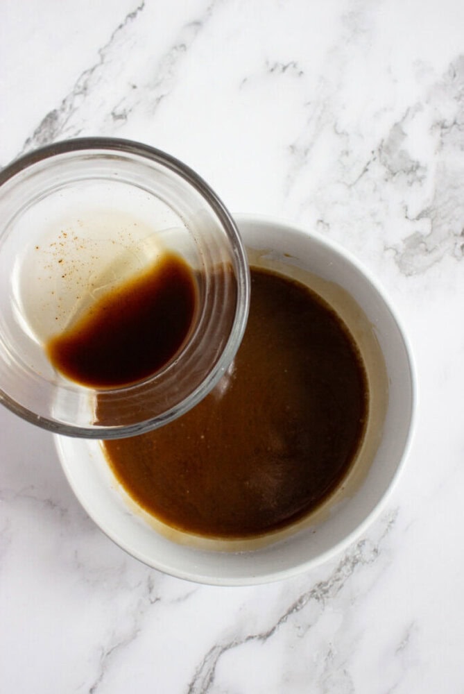 A glass cup with dark brown liquid being poured into a white bowl containing a similar dark brown mixture, on a white marble surface.