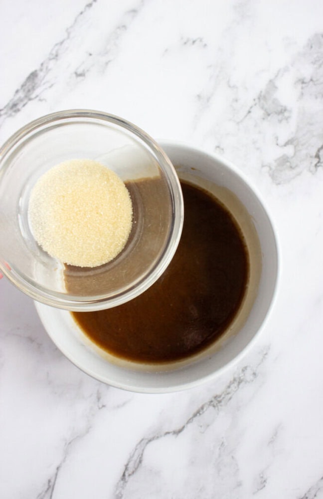 A small bowl of granulated gelatin is being poured into a larger bowl containing a dark brown liquid on a marble surface.