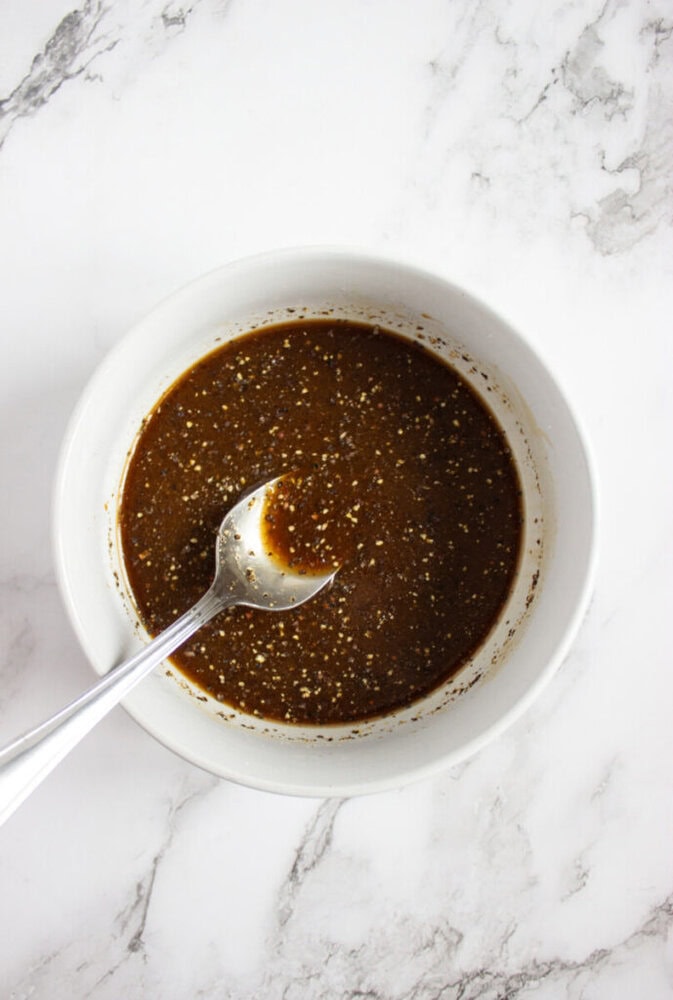A white bowl containing a dark brown liquid marinade with visible spices, and a metal spoon resting inside, on a white marble surface.