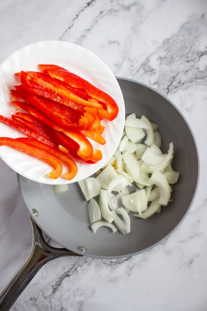 A plate with sliced red bell peppers is being added to a frying pan containing chopped onions on a marble countertop.