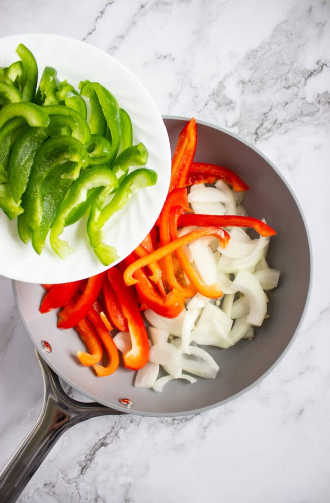 Sliced green peppers are being added from a plate into a pan containing sliced red peppers and onions on a marble countertop.