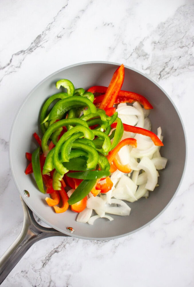 A frying pan with sliced green bell peppers, red bell peppers, and onions on a marble countertop.