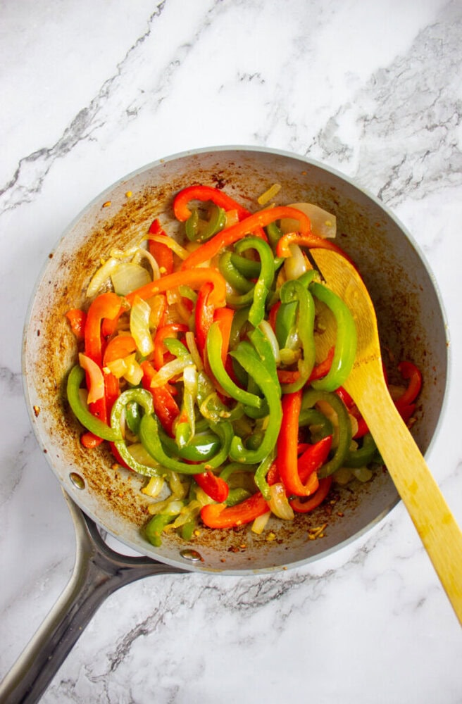 Sliced red and green bell peppers with onions being sautéed in a frying pan, stirred with a wooden spoon on a marble countertop.