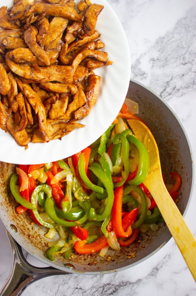 A plate of cooked chicken strips is being added to a pan of sautéed green and red bell peppers and onions on a marble countertop.