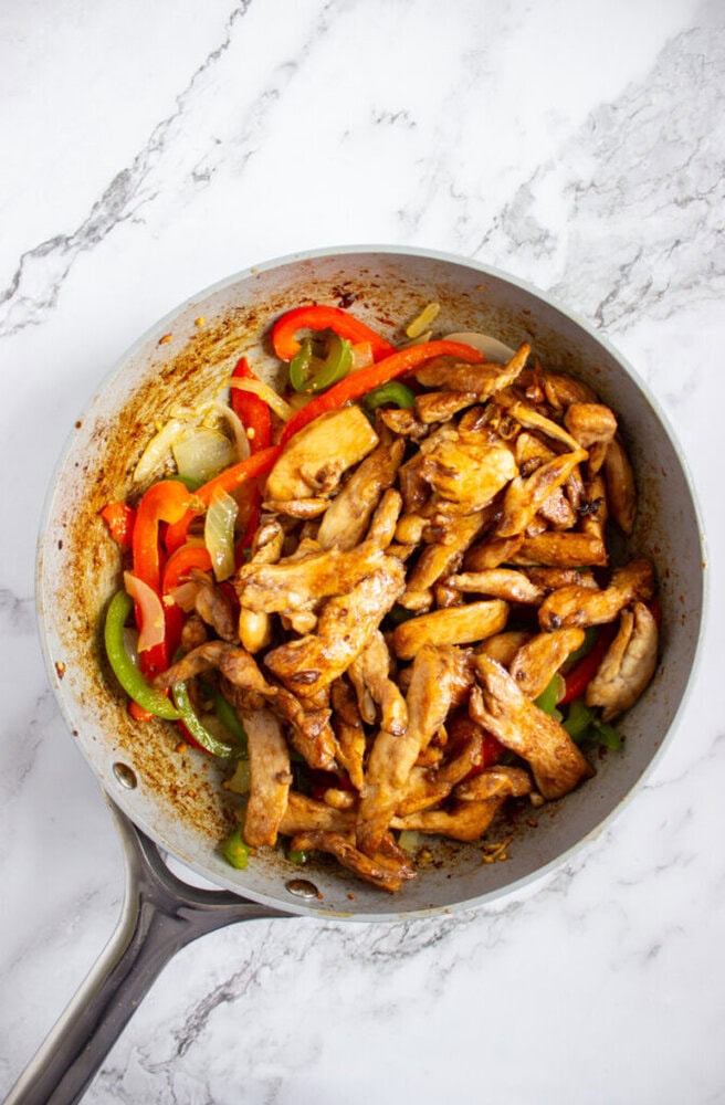 A frying pan with cooked strips of chicken, red and green bell peppers, and onions on a marble countertop.