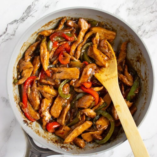 A skillet filled with cooked chicken strips and sliced red and green bell peppers in a brown sauce, with a wooden spoon resting inside.