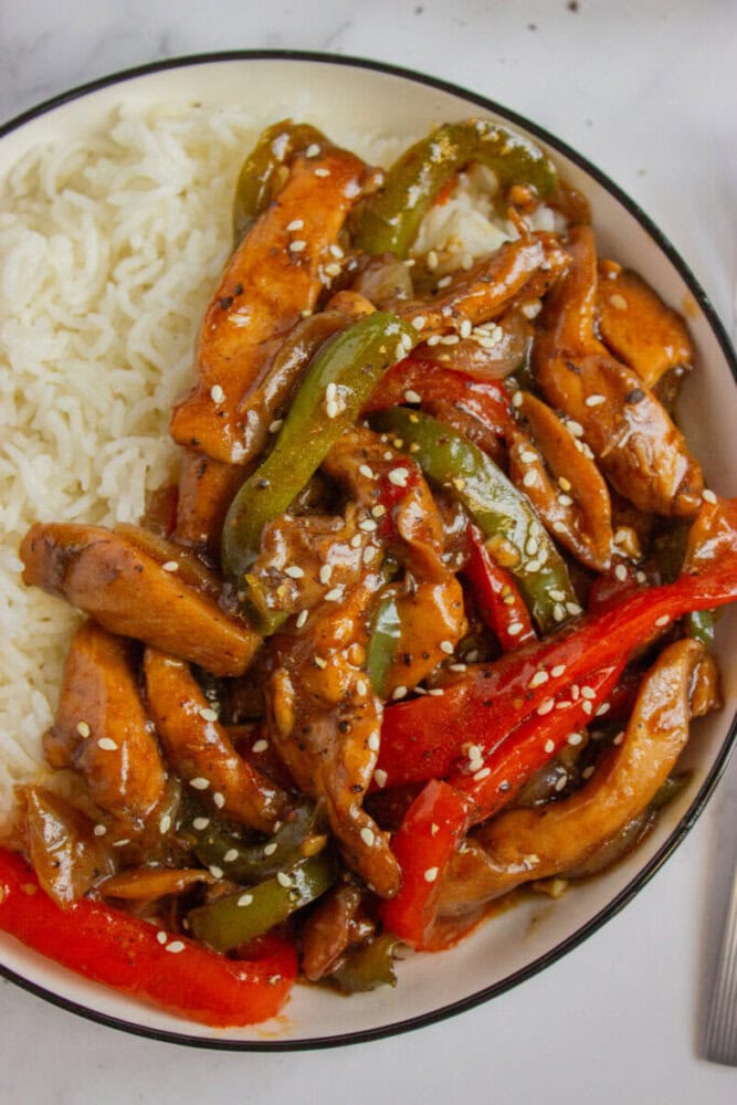 A bowl of white rice topped with stir-fried chicken, red and green bell peppers, and sesame seeds in a brown sauce.