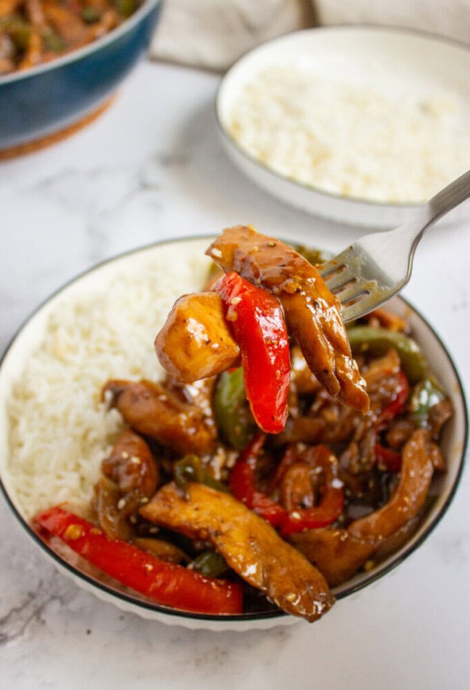 A bowl of white rice and stir-fried chicken with red and green bell peppers; a fork holds a bite of chicken and pepper above the bowl.
