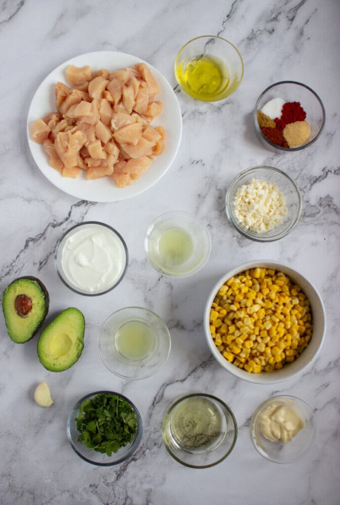 Various ingredients are arranged on a marble surface, including raw diced chicken, corn, avocado, fresh herbs, yogurt, seasonings, oil, lime juice, cheese, and garlic in bowls.