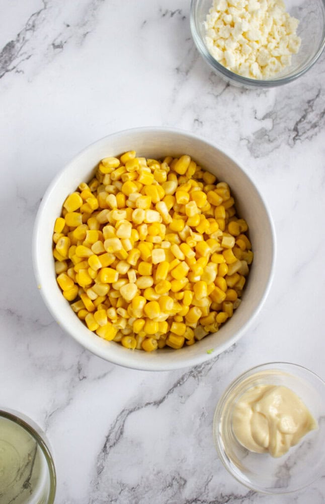 A white bowl filled with corn kernels on a marble surface, surrounded by small bowls containing mayonnaise, crumbled cheese, and another ingredient.