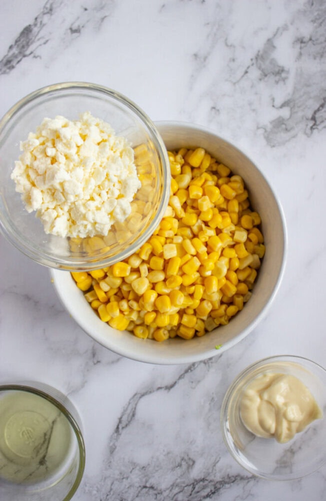 A bowl of corn kernels, a small bowl of crumbled cheese, and separate bowls of mayonnaise and liquid on a marble surface.