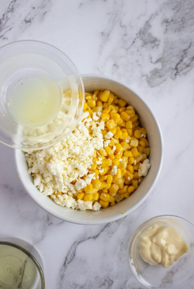 A bowl with corn and crumbled cheese is being topped with a liquid from a small container; other ingredients are in small bowls on the side.