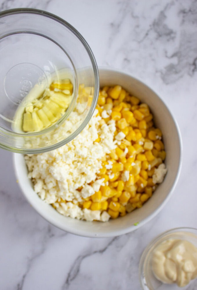 A bowl with corn and crumbled cheese is being topped with melted butter from a glass dish. A small bowl of mayonnaise sits nearby on a marble surface.