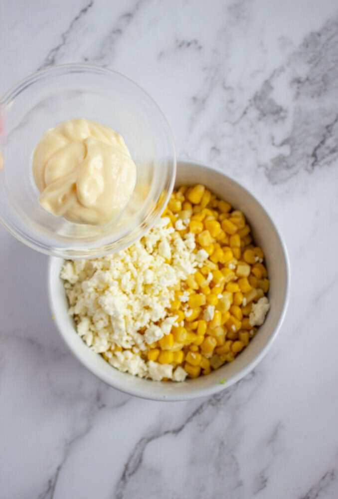 A bowl with corn and crumbled cheese, with mayonnaise being poured in from a small container, on a marble surface.