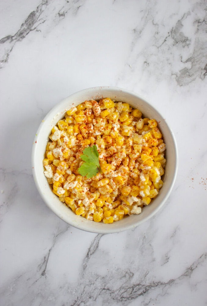 A white bowl filled with corn topped with crumbled cheese, chili powder, and a cilantro leaf, placed on a marble surface.