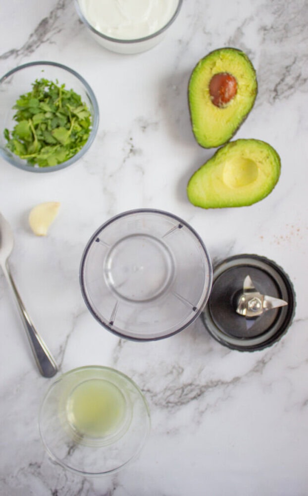 Top-down view of a blender cup with blade, a halved avocado, cilantro, a garlic clove, a spoon, a bowl of yogurt, and a small glass of lime juice on a marble surface.