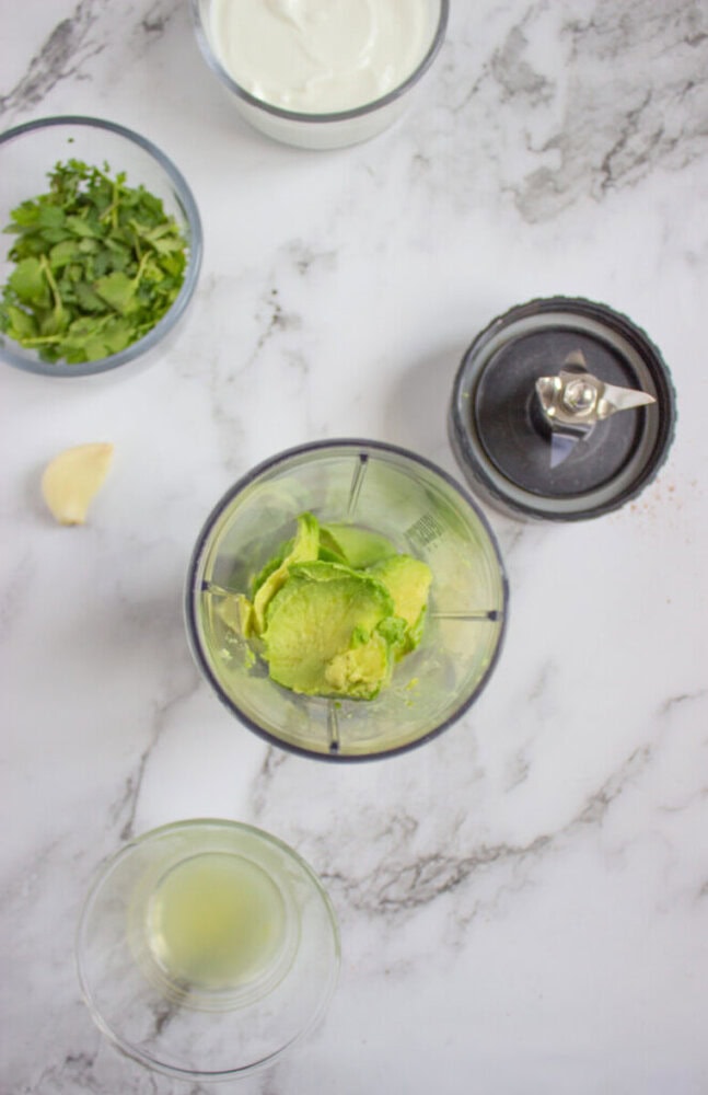 Top-down view of a blender cup with avocado, surrounded by a garlic clove, glass of lime juice, chopped herbs, blender blade, and a bowl of yogurt on a marble surface.