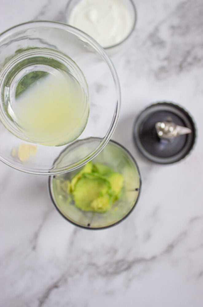 A bowl of lime juice is being poured into a blender containing avocado slices, with a small bowl of garlic and a bowl of yogurt nearby on a marble surface.