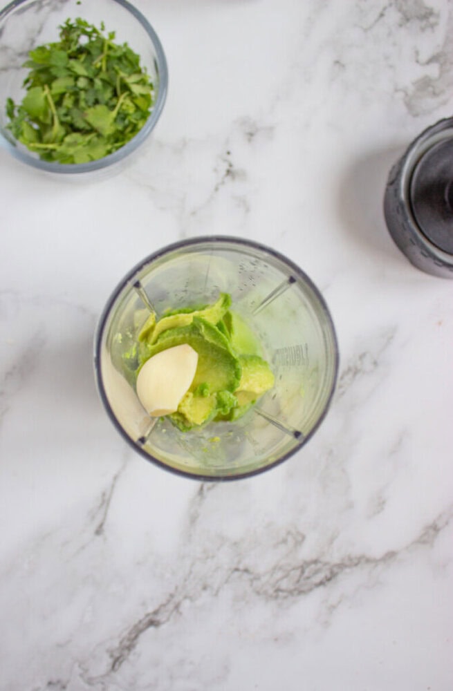 A blender cup with avocado halves and a garlic clove inside, with a bowl of cilantro and a blender lid nearby on a marble countertop.