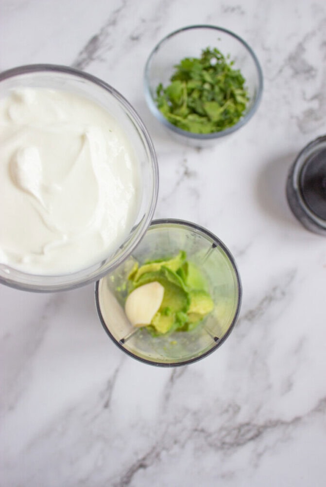 A blender cup with avocado and garlic, a bowl of yogurt, a bowl of chopped herbs, and a pepper grinder on a marble surface.