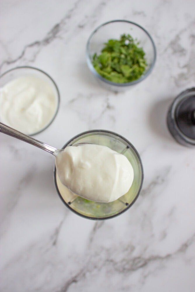 A spoonful of yogurt is held over a blender cup, with bowls of chopped herbs and more yogurt in the background on a marble countertop.