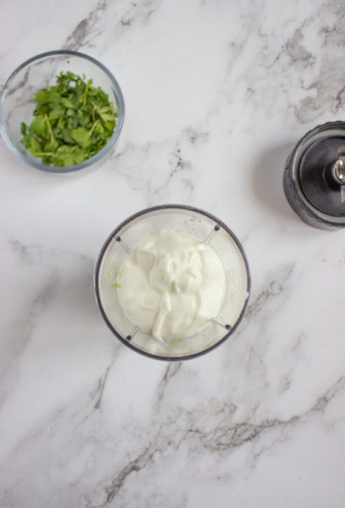 A small blender cup filled with yogurt sits on a marble countertop next to a bowl of chopped cilantro and a black pepper grinder.