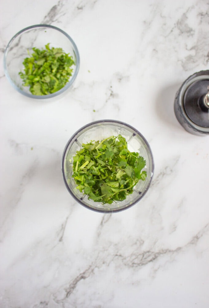 Overhead view of chopped green herbs in two small glass bowls and the top of a blender on a white marble surface.