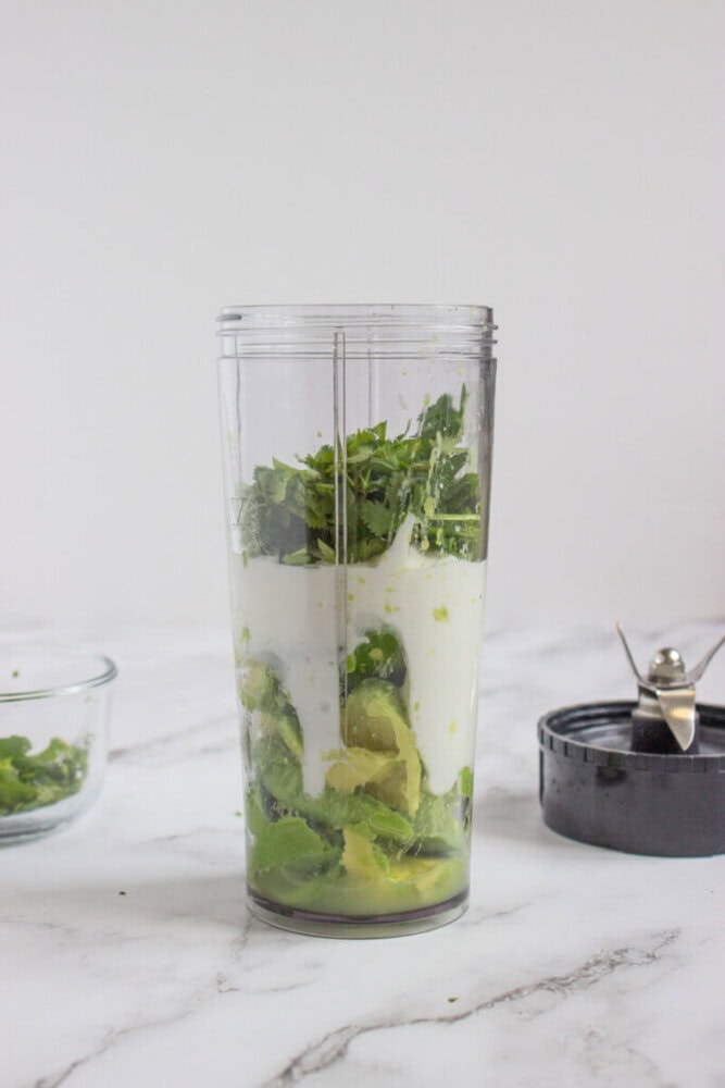 A blender cup filled with avocado, yogurt, leafy greens, and herbs sits on a marble counter next to a blade attachment and a small glass bowl.
