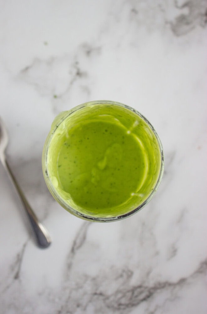 A glass jar filled with green sauce or dressing sits on a white marble surface next to a metal spoon.