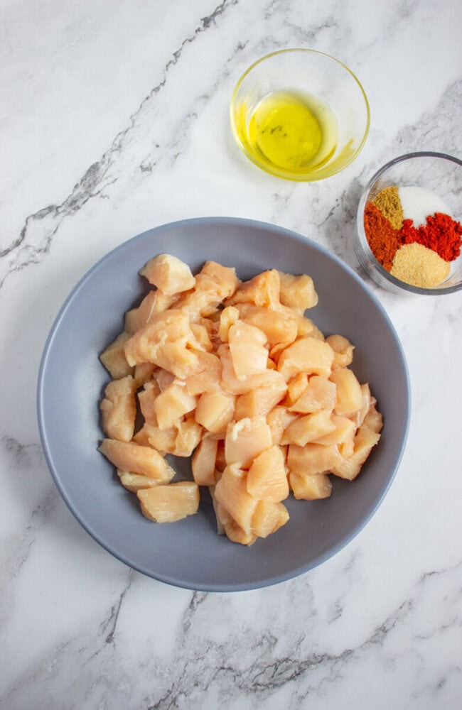 A bowl of raw diced chicken breast, a small bowl of olive oil, and a bowl of assorted spices on a marble countertop.