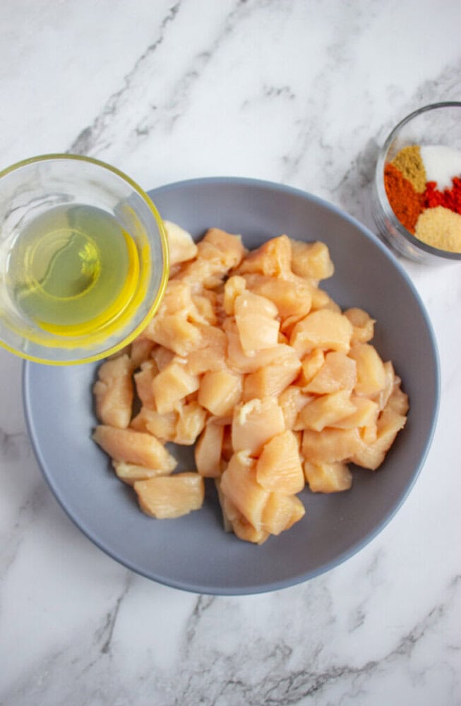 A bowl of raw, diced chicken breast with a small glass bowl of oil and a small bowl of assorted spices on a marble surface.