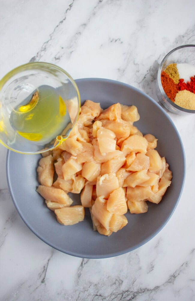 A bowl of raw, cubed chicken breast with oil being poured over it, next to a small bowl containing various spices on a marble countertop.
