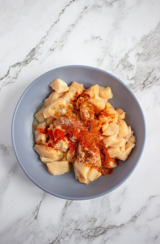 A gray bowl filled with raw, cubed chicken breast topped with various powdered spices sits on a white marble surface.