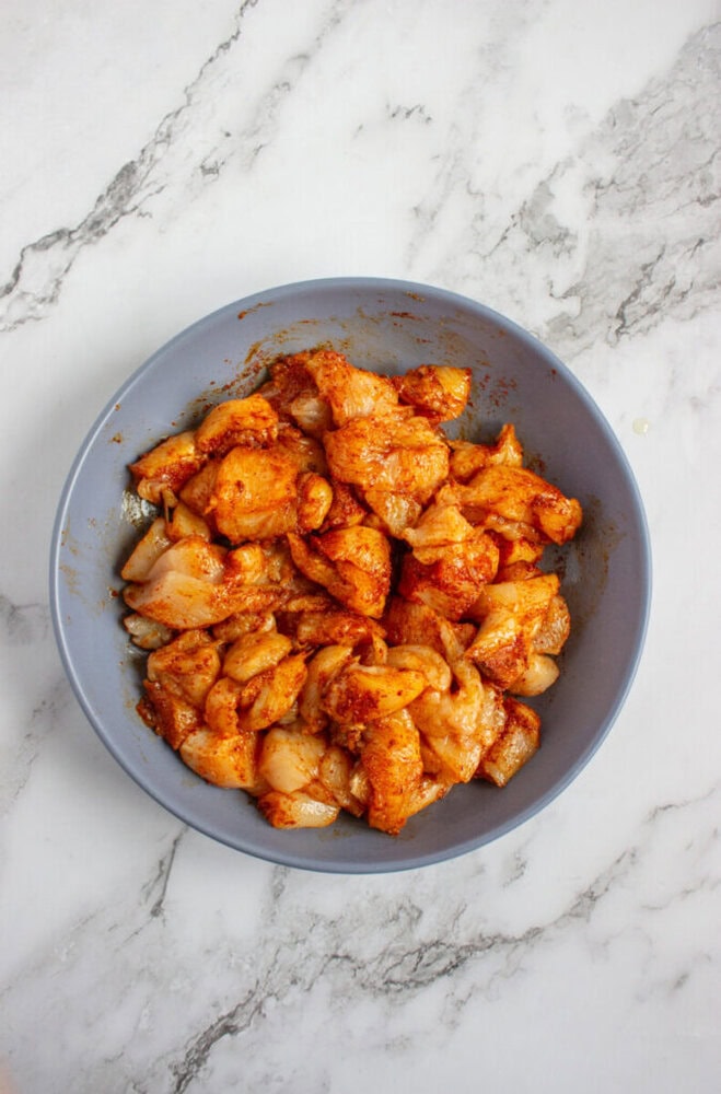 A bowl filled with raw, diced chicken pieces coated in a reddish-orange spice mixture on a marble countertop.