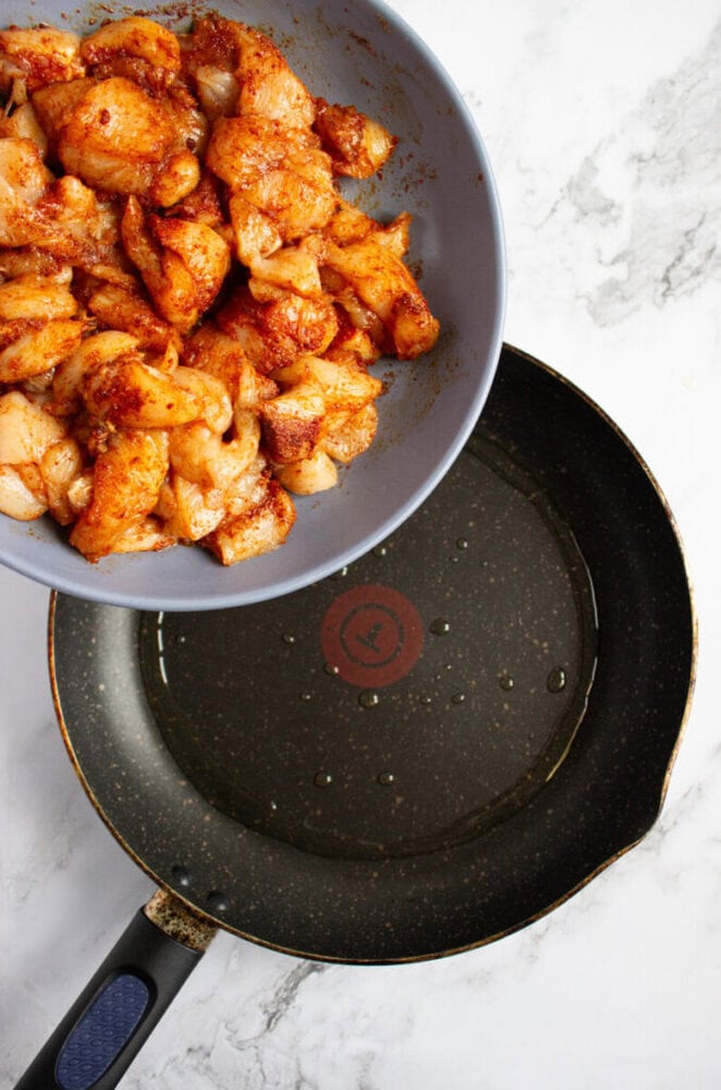A bowl of seasoned raw chicken pieces held above a frying pan with a small amount of oil heating on a stovetop.