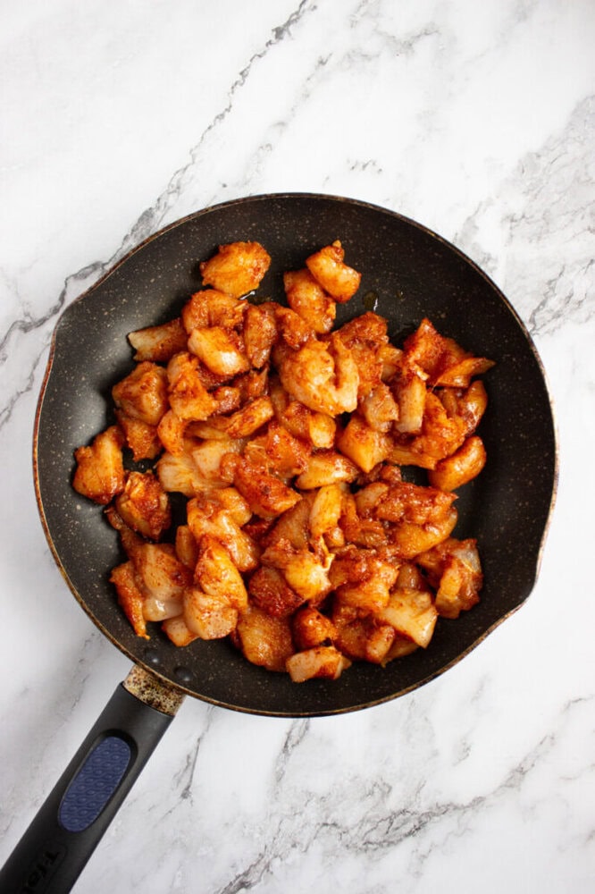 A frying pan containing cooked, seasoned chicken pieces sits on a white marble surface.