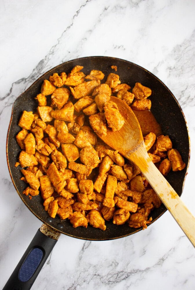 A frying pan filled with seasoned, cooked chicken pieces being stirred with a wooden spoon on a marble countertop.